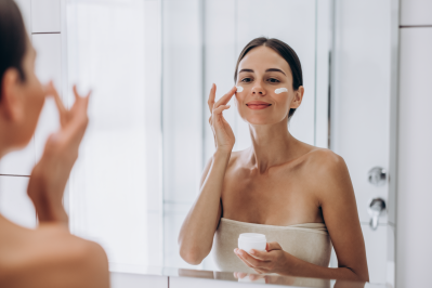 Woman applying face cream while looking in a mirror in a clean, minimal bathroom setting.